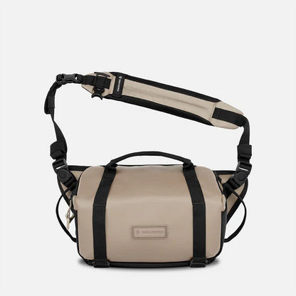 A beige and black messenger bag with a top handle, adjustable shoulder strap, and a small rectangular brand patch on the front, shown against a light gray background.