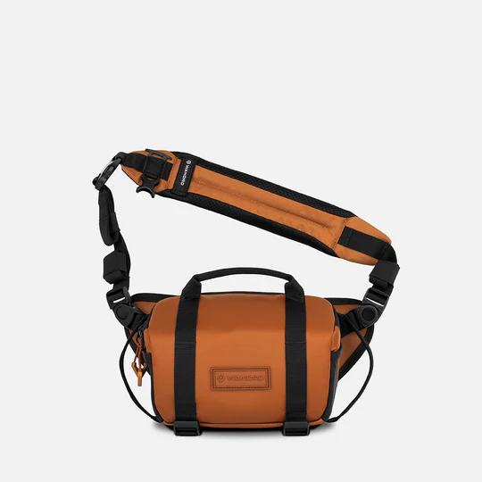 A Wandrd crossbody bag in a warm orange-brown color with black straps and trim, shown against a light gray background.