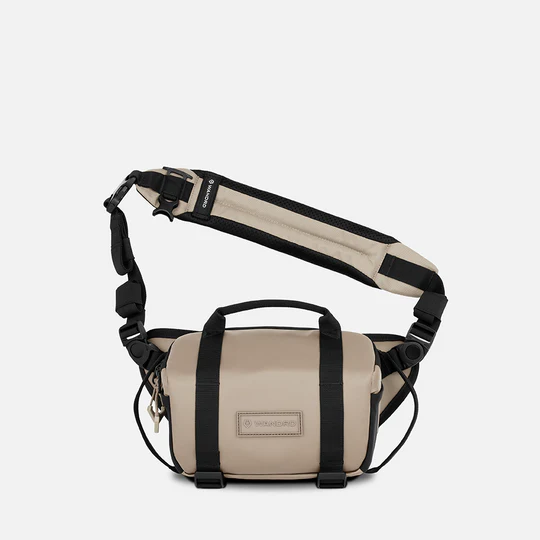 A beige and black Wandrd brand crossbody bag with an adjustable shoulder strap, top handle, and front buckle closure, shown against a light gray background.