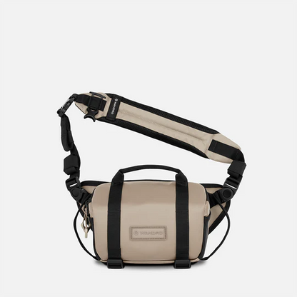A beige and black Wandrd brand crossbody bag with an adjustable shoulder strap, top handle, and front buckle closure, shown against a light gray background.